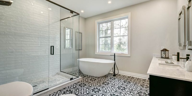 A contemporary bathroom featuring a glass-enclosed shower, a freestanding bathtub, and stylish patterned floor tiles, complemented by a large window.