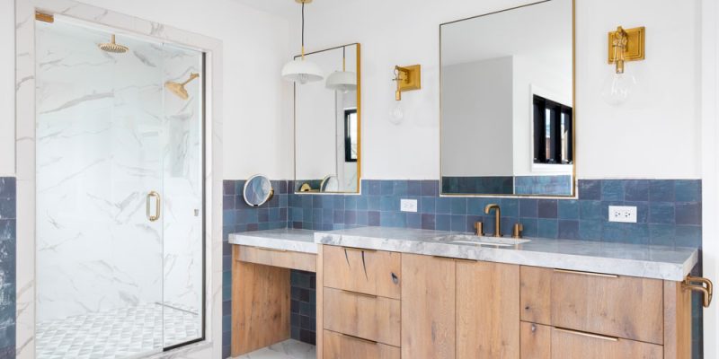 A chic bathroom design with dual sinks, marble countertops, wooden cabinetry, and gold accents, paired with a glass-enclosed shower and modern lighting.
