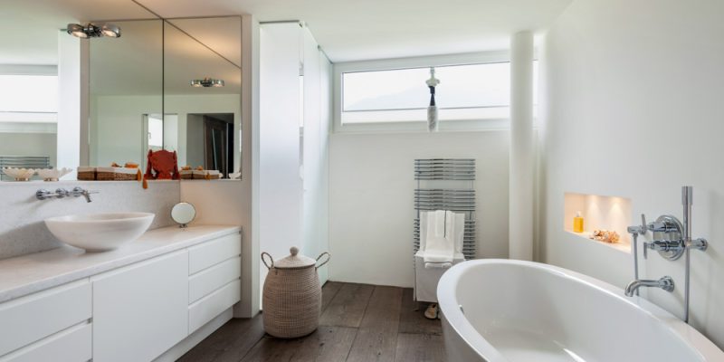 A sleek, minimalist bathroom design with a freestanding bathtub, white vanity, large mirror, and natural light streaming in through a high window.