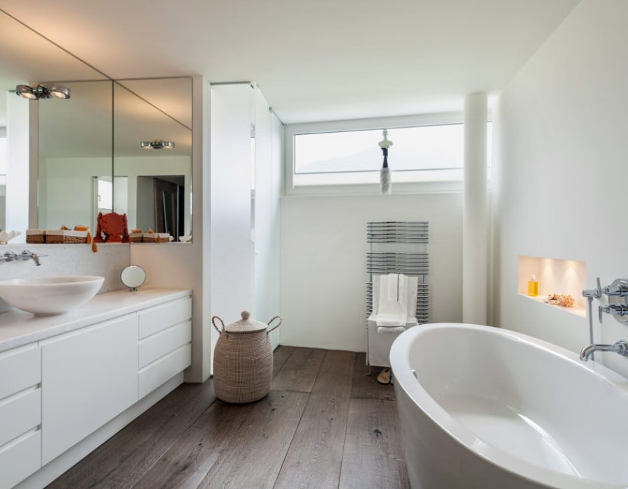 A sleek, minimalist bathroom design with a freestanding bathtub, white vanity, large mirror, and natural light streaming in through a high window.