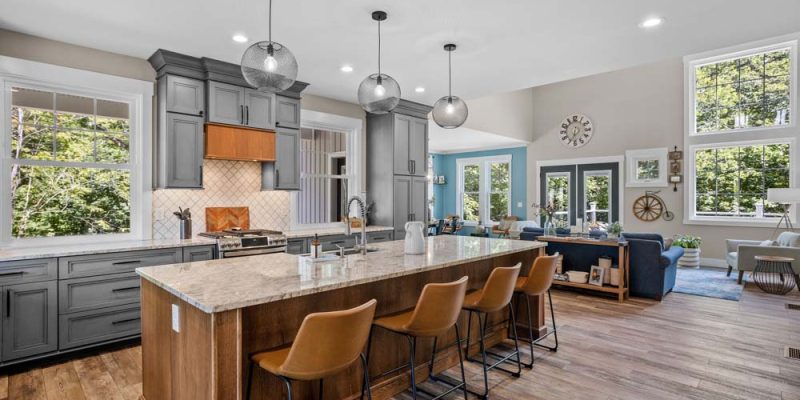 A modern kitchen featuring dark gray cabinets, stainless steel appliances, and a large island with seating, seamlessly connected to a bright living area with large windows and stylish furnishings.