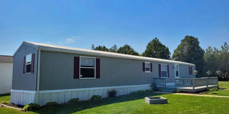 An updated mobile home exterior painted in soft gray with a white-trimmed porch and red window frames, set in a neatly manicured lawn for a homely appeal.