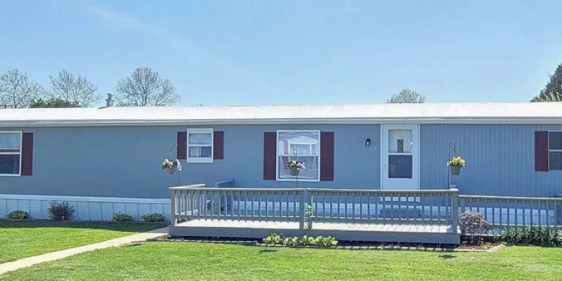An elegantly remodeled mobile home featuring a new gray facade and red window accents, complemented by a welcoming front porch with railing and potted plants.